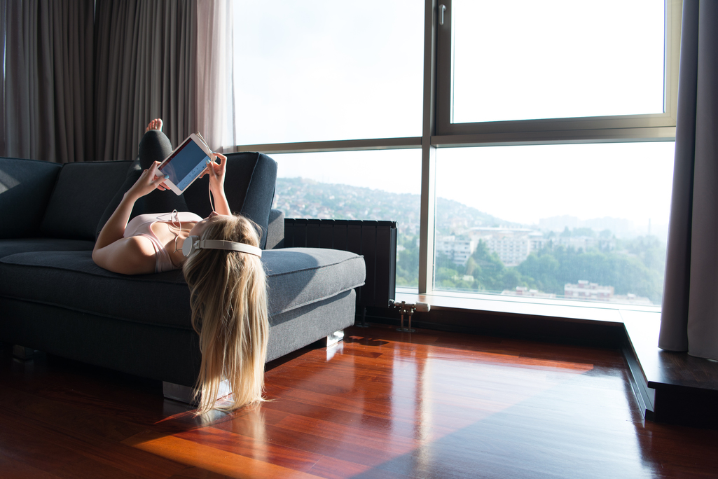 Woman laying on a couch with headphones on in front large windows looking out over a city.