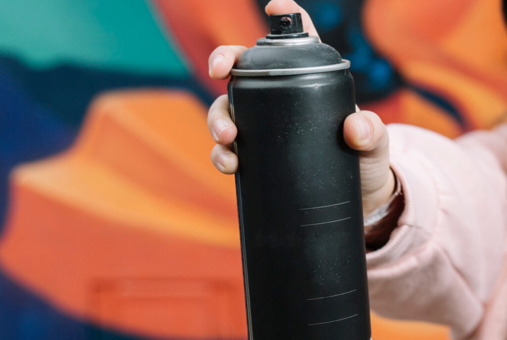 Someone holding up a black spray paint can with graffiti on a wall behind them.